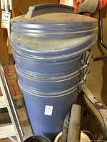 Three large blue Rubbermaid plastic garbage cans with lids showing wear and dust.