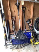 Full view of the assortment of garden tools including leaf rake, multiple snow shovels, and a chrome shovel all showing signs of wear