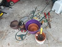 Photo showing assortment of garden tools and pots including purple pot nest with smaller terracotta pots, cream pot with garden fork, brown pots, red handle shears, green cultivator, garden knife, and several plant support stakes.