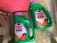 Two unopened green Miracle-Gro Shake 'n Feed Ultra Bloom fertilizer containers with purple caps, alongside red gas can and other items on floor.