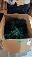 Large open cardboard box filled with green artificial garland strands with embedded lights.