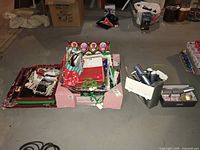 Photo taken from a distance showing the entire lot with boxes and bins containing Christmas wraps, bags, crackers, and ribbons on a basement concrete floor.