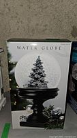 Front side of original box with image of water globe featuring snow-covered Christmas tree on black pedestal base