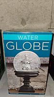 Photo of the water globe in its original blue box depicting the snowman inside and the pedestal base.