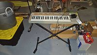 Full view of the keyboard on black X-stand with music holder, connected foot pedal, cables visible, placed in basement setting surrounded by storage items.