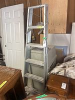 Full view of the metal step ladder leaning against a large plastic container, showing six steps and top platform with metal frame.