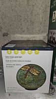 Image of the original box with product picture and text in three languages indicating 'Solar mosaic globe light'. The mosaic globe light features a dragonfly design on a green-blue mosaic sphere.