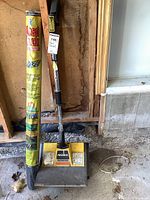 Photo showing Noma Sno Shovel and unopened weed block roll leaning against an unfinished wall in a garage or storage area.