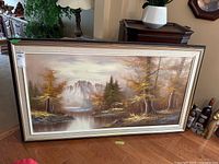 Full view of the framed oil painting showing the landscape scene with lake, trees, mountains, and cabin, placed on the floor against a wall.