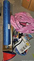 Blue foam pool roller approx 35 inches with Pathfinder pink inflatable raft and assorted pool accessories including beach balls and test kit parts in a cardboard box on concrete floor.