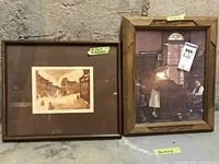 Two framed prints leaning against a concrete wall on the floor, one showing a sepia street scene, the other showing children indoors and signed.