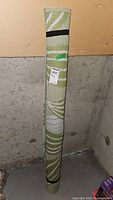 Photo of Sonoma green and white woven rug rolled upright against wall with black strap securing it