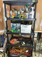 Full view of black plastic shelving unit with five shelves containing various items such as spray cans, boxes, an orange tool, and extension cords. The shelving unit is worn and dusty with no visible damage to structure.