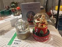 Photo showing two snow globes side by side. The Coca-Cola Santa musical globe is red base with Christmas decorations and a Santa figure holding a bottle. The other globe shows a winter ice skating scene with a white base.