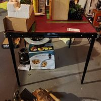 Side view of the folding card table showing red vinyl top and black metal foldable legs against basement background with boxes and other items.