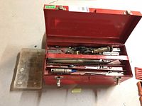 Red metal toolbox opened showing contents of hand tools including wrenches, screwdrivers and socket wrench pieces arranged inside compartments.
