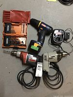 Photo showing three drills (two corded drills and one cordless) with two sets of drill bits laid out on floor.