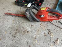 Side angle showing the orange and black Black & Decker electric hedge trimmer with 20-inch blade, placed on concrete flooring with dirt and debris.