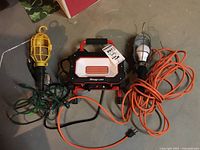 Photo showing three work lights on the floor: Snap-on red and black LED light in center, yellow cage light left, silver cage light right.