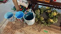 Photo showing two plastic buckets with colorful patterns, a fabric basket with a blue polka dot pattern, and a faux flower wreath placed on a carpeted floor near a wooden table leg.
