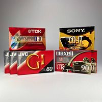 Seven sealed blank audio cassette tapes arranged side by side. Brands visible are TDK, Sony, JVC GI, and Maxell.