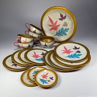 Full set of 18 vintage tin litho toy dishes arranged, showing various sizes of plates, saucers, coasters, teacups, cream and sugar pieces with colorful leaf litho designs and gold rims.