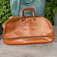Front view of vintage brown leather travel bag showing leather-wrapped handles, brass hardware, zipper on bottom compartment for shoes.