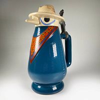Side angle view of the ceramic beer stein showing the blue body, orange sash detail, and cream-colored farmer hat lid with painted features.