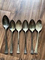 Six spoons laid out on wooden floor, showing plain handle design and tarnishing.