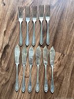 Top view image showing 5 forks and 6 butter knives arranged vertically on wooden surface. Handles have tarnish and wear marks.