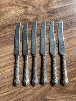 Set of six silver-plated tea cutters with ornate floral and scrollwork handles placed on a wooden surface.