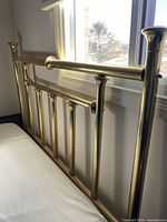 Brass headboard positioned against a wall near window with visible horizontal and vertical rods.
