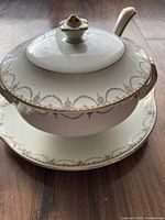 Photo showing closed tureen with lid and gold scalloped and dotted decorative pattern, with underplate underneath on wood floor.
