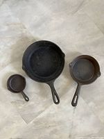 Three cast iron skillets of different sizes shown on floor, all with pour spouts and signs of rust.