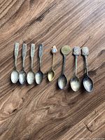 Front view of eight collectible spoons showing different designs and sizes, including vintage engraved spoons and souvenir spoons with emblems.