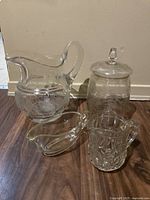 Four clear glass items: one large heavy jug with floral etching, two small creamers, and one lidded jar on wooden floor against cream wall.