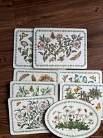 Seven ceramic trivets and place mats with botanical flower and butterfly designs and green leaf borders, arranged on table