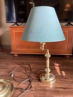 Full view of table lamp with blue-green shade, polished brass base and column, with power cord visible on wood floor.