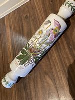 Full view of the ceramic rolling pin showing detailed botanical floral design in multiple colors along the cylindrical body with leafy patterns on the handles.