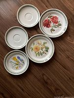 Top down photo of five ceramic saucers and side plates with botanical floral patterns and green leaf border on wooden surface. Two plates are plain with green leaf border and three plates show different flowers.