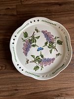 Top view of the white ceramic fluted platter with purple lilac flowers, green leaves, and butterflies, showing naturalistic botanical artwork and fluted rim with cutout details.