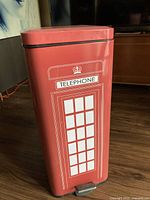 Front view of the red metal trash bin resembling a British telephone booth, showing rectangular shape, red color, white window graphics, and foot pedal.