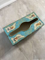 Top angle view of turquoise tin tissue/dryer sheet holder showing floral decals and dispensing slot.
