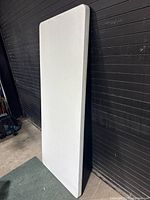 Front view of the white folding table in folded position showing the top surface. The table is rectangular and about 6 feet long.