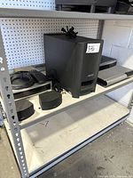 Photo showing a large Bose subwoofer speaker, a media unit, two satellite speakers, a remote control, and cables on a metal shelving unit.