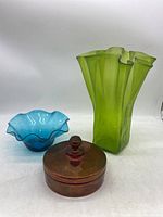 Three colored glass items on a white background: cranberry round covered dish with lid, blue scalloped bowl, and tall green wavy rim vase.