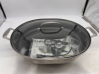 Top view of stainless steel electric skillet with glass lid, showing inside with sealed plastic bag containing power cord and manual.