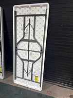 White 6 ft plastic folding table shown folded on its side with metal folding legs and support frame visible on the underside.