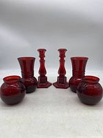 Front view of 5 red glass items including 3 vases and 2 candlestick holders arranged in a line on a white background.
