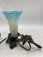 Side view showing blue and white fluted glass floral shade and metal leaf base with bird design, power cord and plug visible.